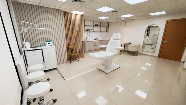 Sala de procedimentos da Dermalink Academy com maca elétrica branca, mochos, carrinho auxiliar dourado e espelho em arco
