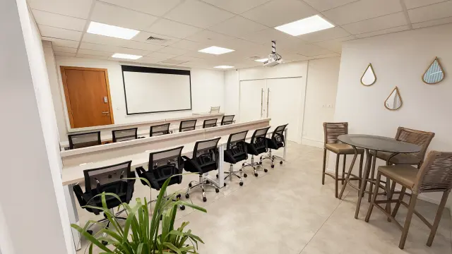 Sala de treinamento com bancadas para 16 pessoas, cadeiras giratórias, projetor, tela de projeção e mesa alta de apoio